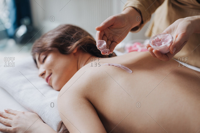 A masseuse putting natural moisturizing cosmetic cream on woman's back at spa salon.