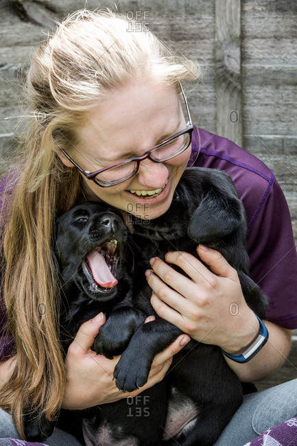Blond woman wearing glasses hugging two Black Labrador puppies.