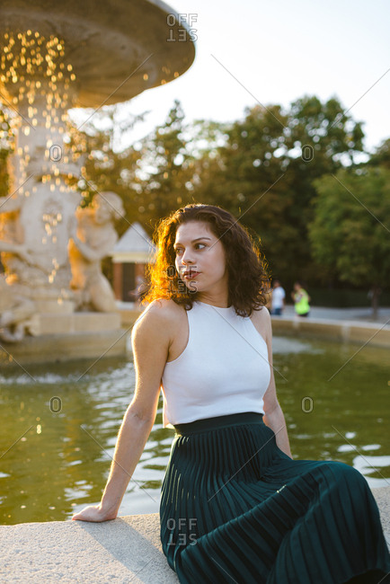 Beautiful lady in tank top and green skirt looking away seated on fountain ledge at sunset