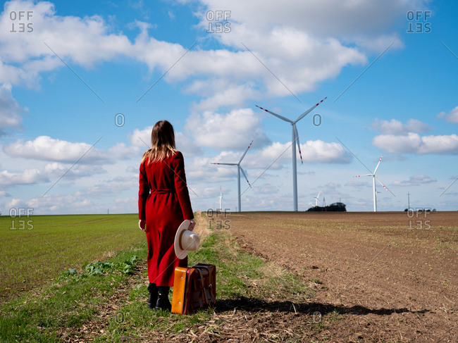 Woman in red coat with suitcase on field with wind turbines