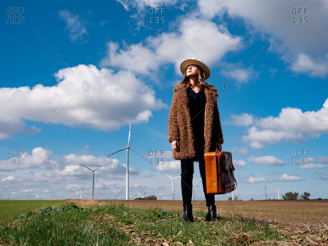 Woman in coat with suitcase on field with wind turbines