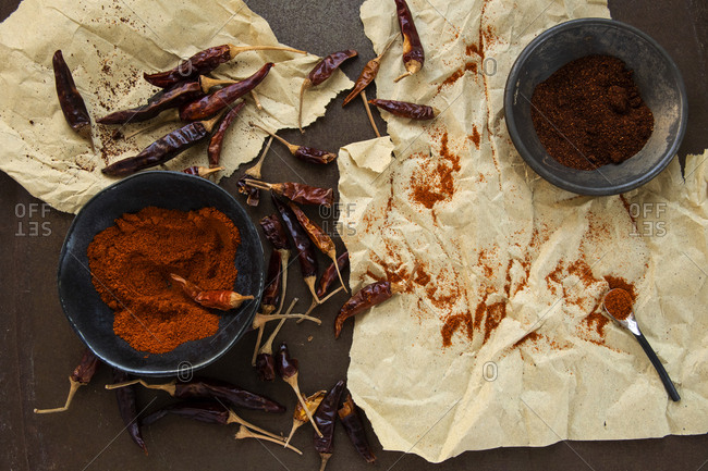 Cayenne and paprika powder in decorative antique bowls circled by dried peppers on parchment paper