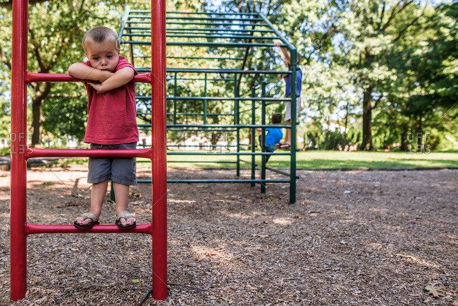 Toddler boy looking sad on playground ladder with older boys playing