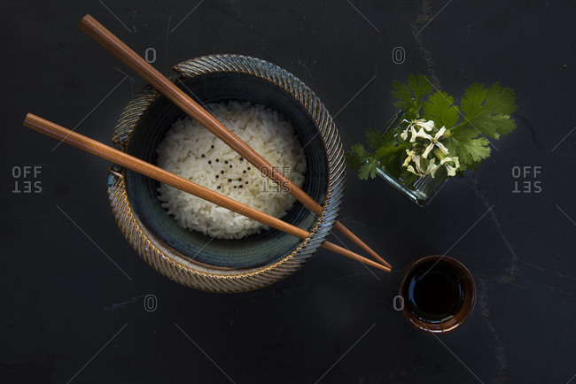 An Asian-style bowl of rice with chopsticks, soy sauce, herb on black