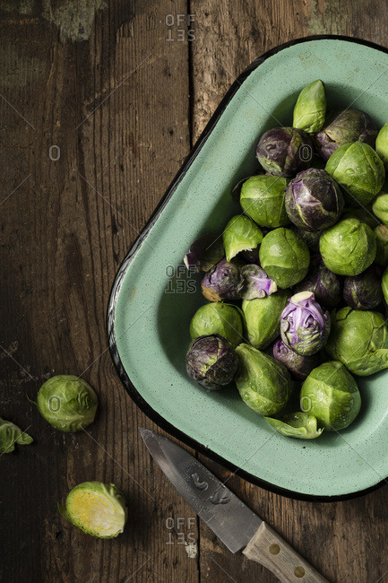 Raw Brussel sprouts in a vintage, green enamel dish, on a rustic wooden surface. A knife is alongside.