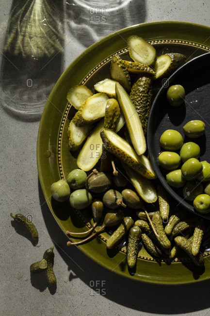 A selection of pickled gherkins, caper berries and green olives on an olive green plate. Shadows on the concrete background suggest glasses of wine.