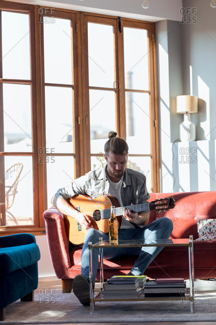 Shot of hipster young man playing the acoustic guitar while sitting on the couch in the living room at home.