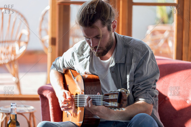 Shot of hipster young man playing the acoustic guitar while sitting on the couch in the living room at home.