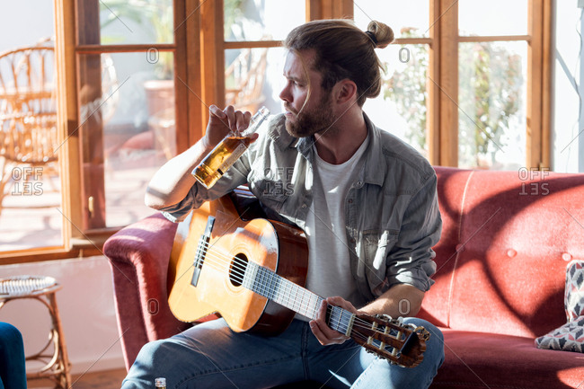 Shot of hipster young man drinking beer while playing the acoustic guitar and sitting on the couch in the living room at home.