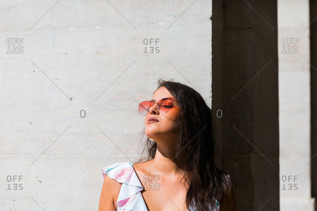 Peaceful gorgeous woman in trendy outfit and shiny sunglasses standing on white wall on scenic street