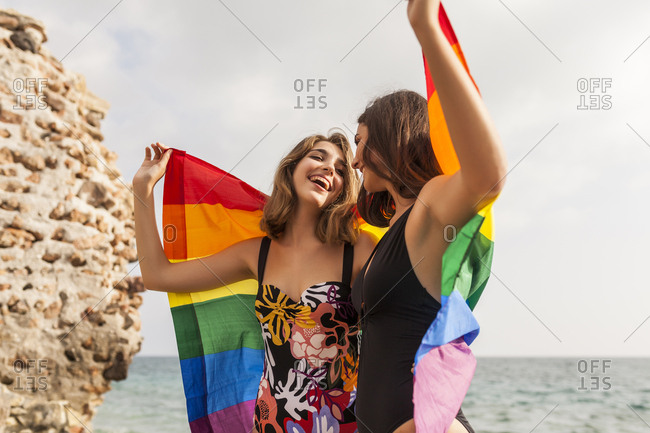 Lesbian couple standing on beach hugging wrapped on colorful flag of LGBT movement during summer vacation looking at each other