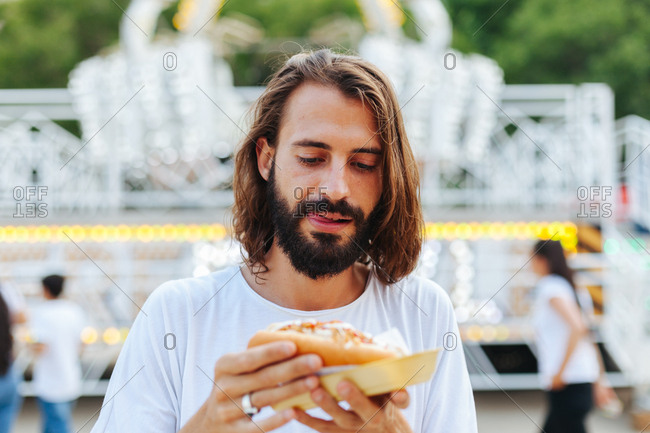 Delighted bearded man biting sandwich with closed eyes while standing beside lighted attraction at carnival