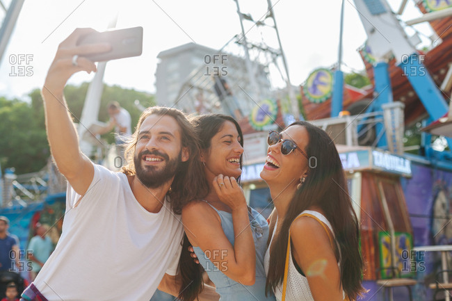 Cheerful smiling tanned people taking photo on smartphone while standing next to attraction at carnival