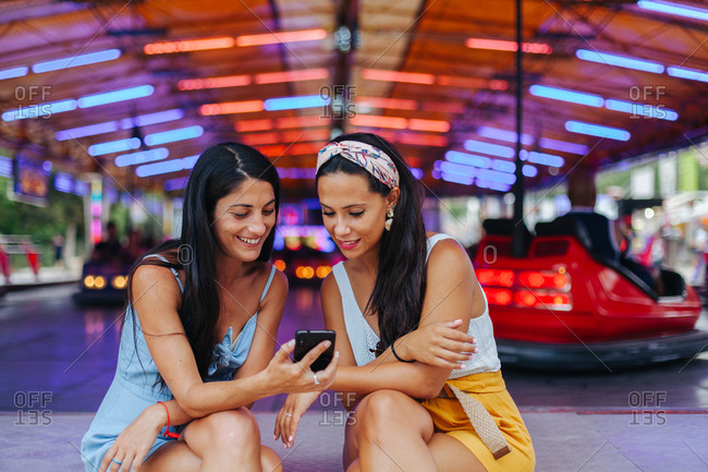 Content smiling tanned women taking photo on smartphone while sitting next to amusement attraction at carnival