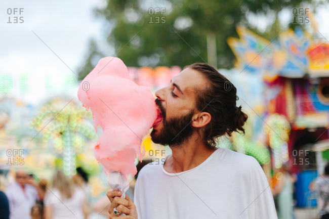 Side view of charming smiling brunette biting cotton candy while standing next to multicolored attraction with neon lights at funfair