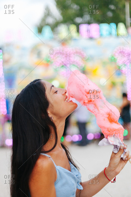 Side view of charming smiling brunette biting cotton candy while standing next to multicolored attraction with neon lights at funfair