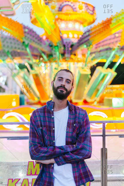 Calm hipster man in casual clothes standing with crossed arms beside lighted colorful carousel at fairground