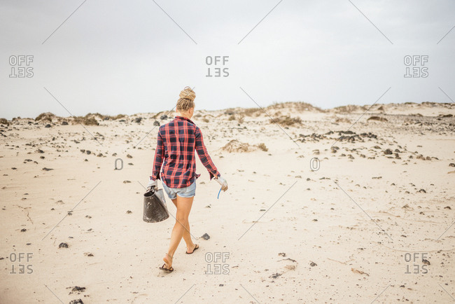 Positive hipster woman in casual clothes and gloves collecting trash into bag while squatting on deserted beach