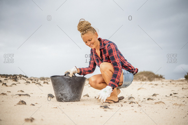 Positive hipster woman in casual clothes and gloves collecting trash into bag while squatting on deserted beach