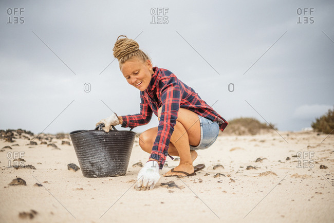 Positive hipster woman in casual clothes and gloves collecting trash into bag while squatting on deserted beach