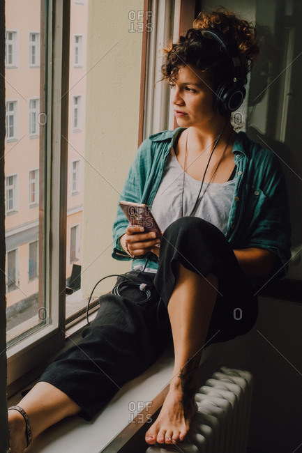 Pensive curly woman in headphones listening to music while browsing smartphone and sitting on window sill in apartment looking out of the window