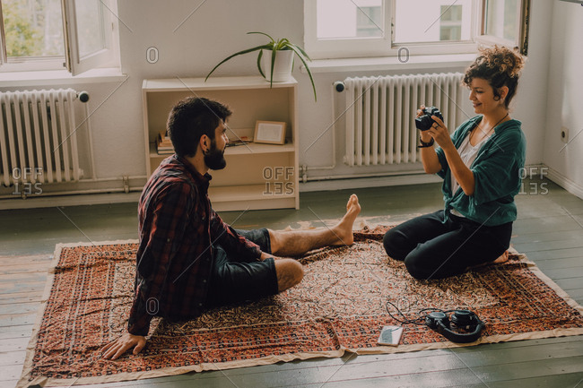 Side view of hipster woman taking picture of man sitting barefoot and posing on apartment floor