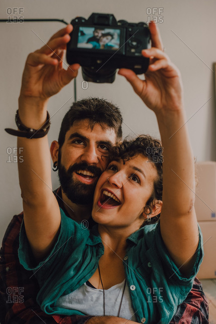 Affectionate hipster couple hugging and taking selfie with photo on camera while sitting on floor at home