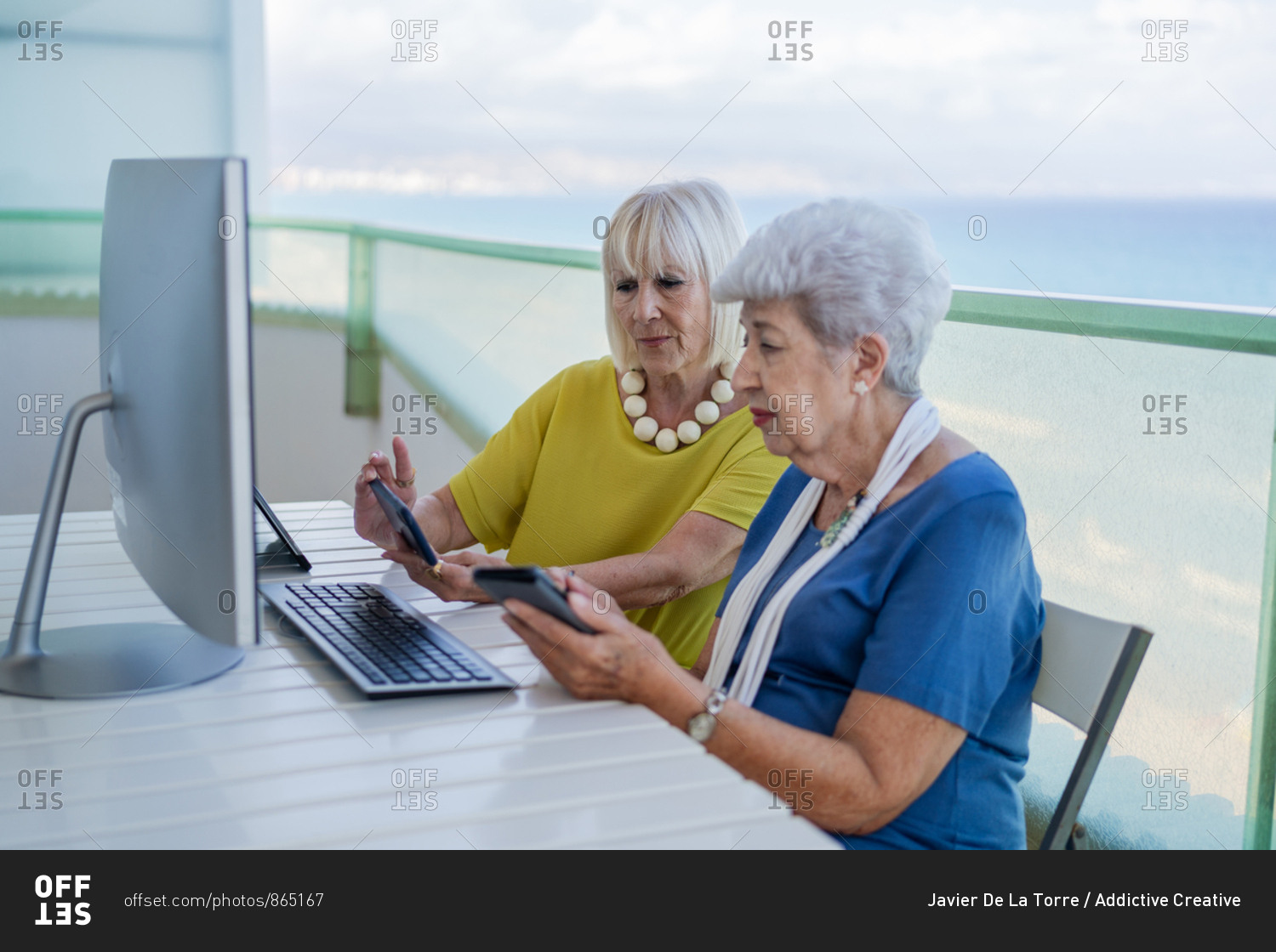 Stylish old friends browsing social media on tablet while sitting at table with computer on balcony on resort