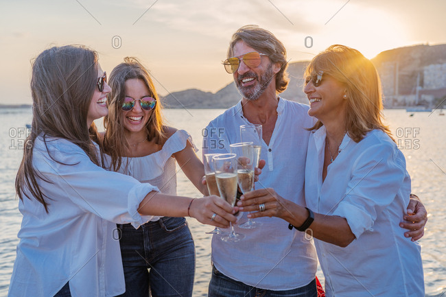 Cheerful mature parents and young daughters clinking glasses of wine and laughing while celebrating family reunion in evening on resort