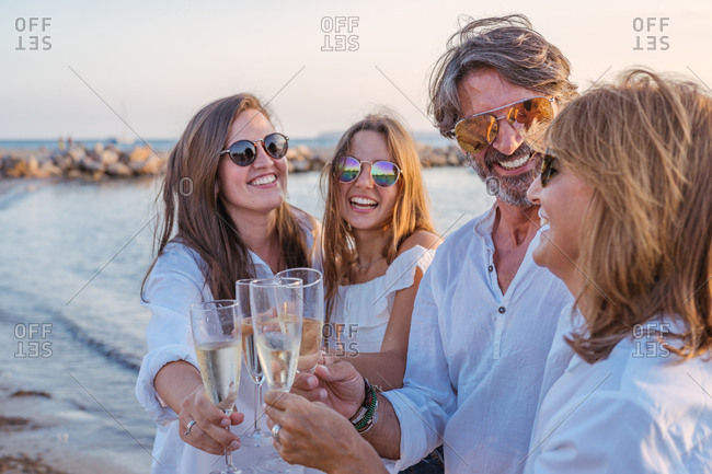 Cheerful mature parents and young daughters clinking glasses of wine and laughing while celebrating family reunion in evening on resort