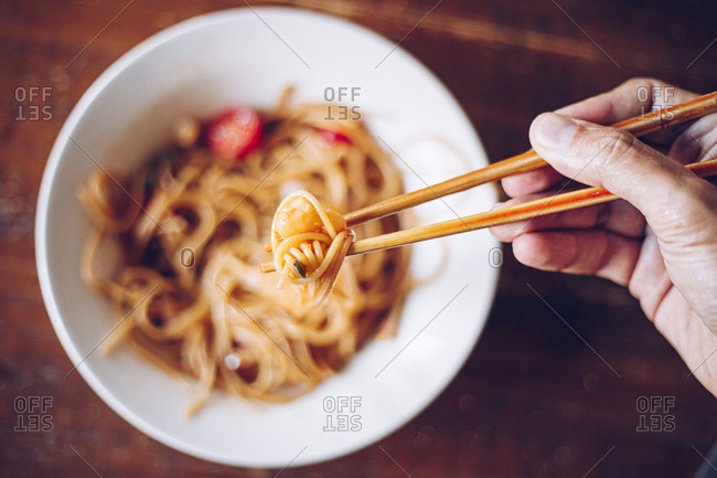 From above soft focus of crop person holding shrimp by wooden chopsticks while enjoying noodles on blurred background