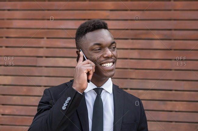 Positive smiling successful African American male speaking on mobile phone and holding finger in pocket while standing beside urban striped wooden wall