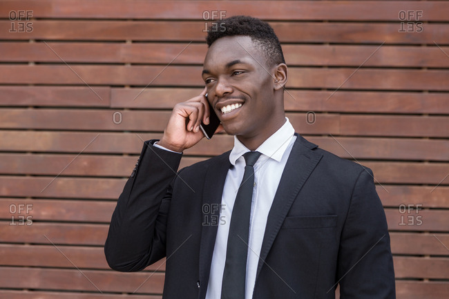 Positive smiling successful African American male speaking on mobile phone and holding finger in pocket while standing beside urban striped wooden wall