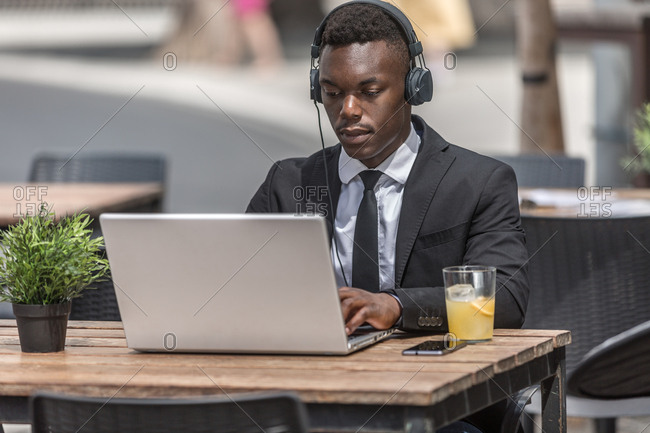 black man in suit sitting in cafe outside with laptop and in wired headphones