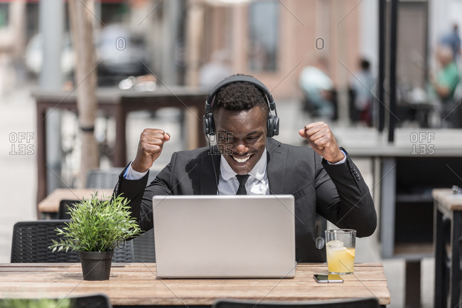 Happy joyful black man in suit rejoicing at success with raised hands sitting in cafe outside with laptop and in wired headphones