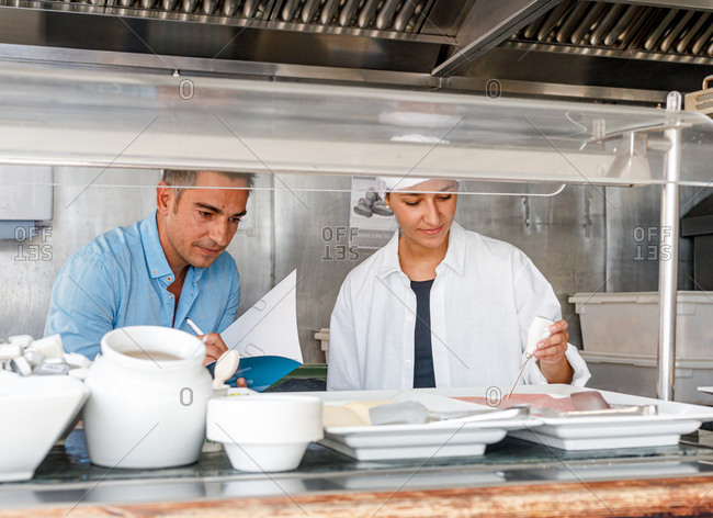 Attentive senior employee writing in notebook results while adult female colleague in sterile clothes using digital device for test food quality in canteen