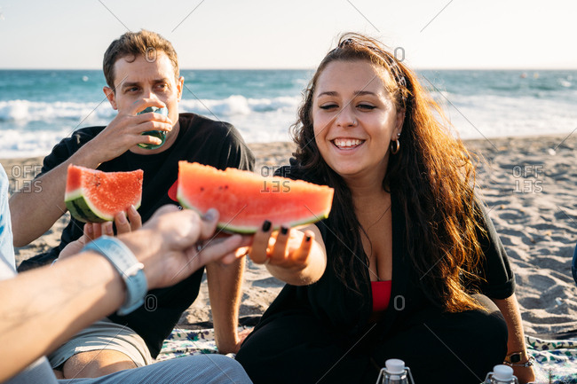 Smiling woman gives a piece of watermelon to her friend with her friend who drinks orange juice on the beach