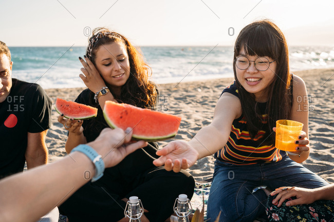 Smiling woman gives a piece of watermelon to her friend with her friend who drinks orange juice on the beach