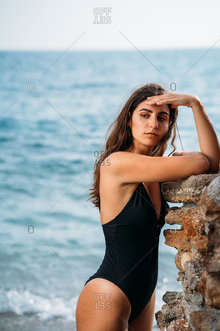 Side view of attractive pensive woman in black swimwear relaxing on seashore leaning by stone wall and looking away