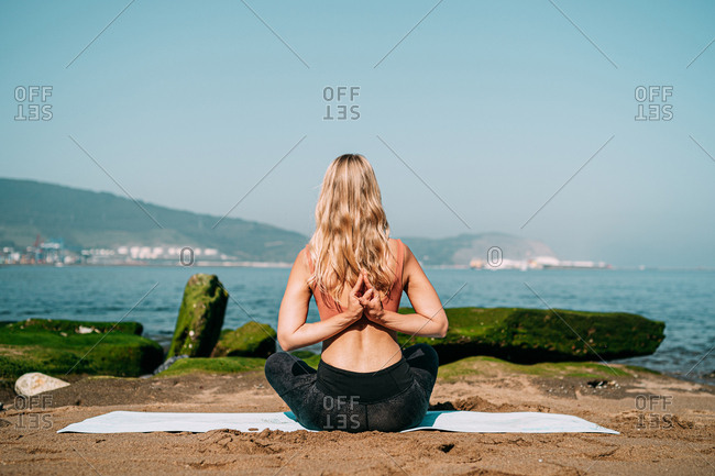 From behind sportive woman practicing yoga asana with hands behind back while sitting on mat at tranquil coast