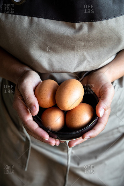 From above crop cook in gray apron standing with fresh chicken eggs in hands for cooking