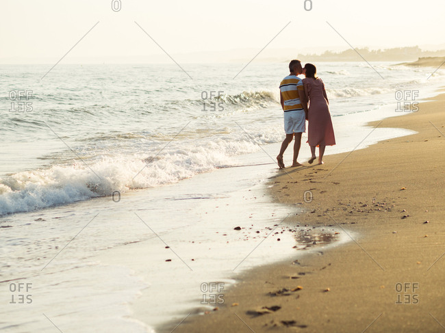 Back view of barefoot man and woman holding hands and carrying shoes while walking on sandy beach towards waving sea on resort