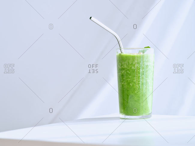 Tall glass of green healthy detox cocktail with metal straw on white minimalistic table over white background