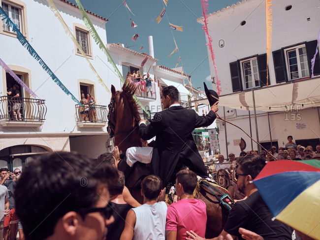 August 25, 2019: Spain, Menorca August 25, 2019: Side view of rider in festive suit sitting on black horse floundering in sandy arena surrounded by surprised joyful people