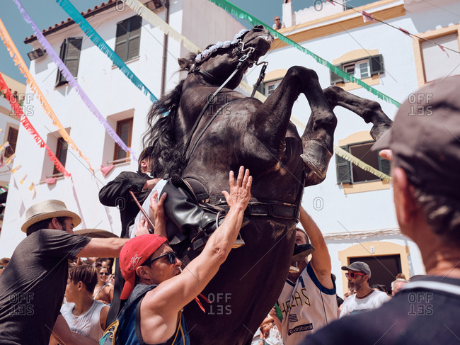 August 25, 2019: Spain, Menorca August 25, 2019: Side view of rider in festive suit sitting on black horse floundering in sandy arena surrounded by surprised joyful people