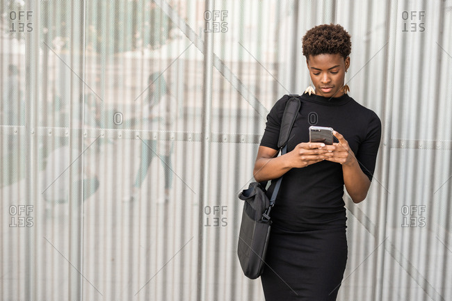 Focused stylish African American woman in black dress messaging smartphone while standing on metal background
