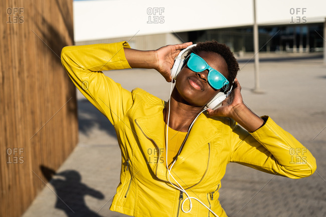 African American woman in stylish bright jacket and bright blue sunglasses using headphones standing near a modern building