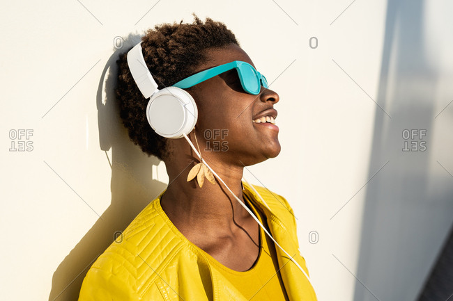 African American woman in stylish bright jacket and bright blue sunglasses using headphones leaning in a white wall