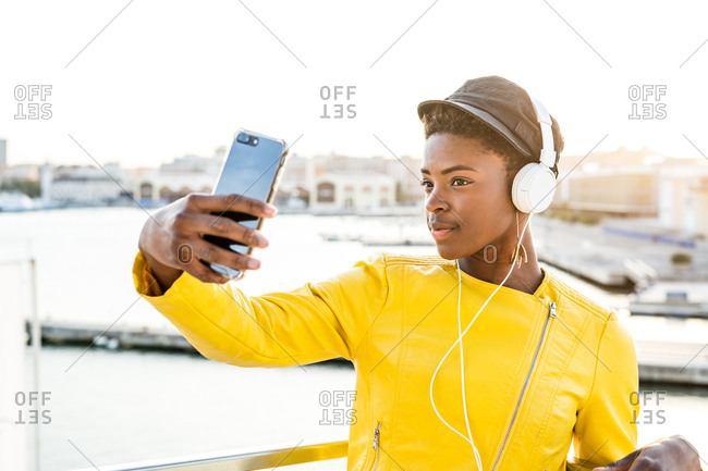 African American woman in stylish bright jacket using mobile phone and listen to music on headphones