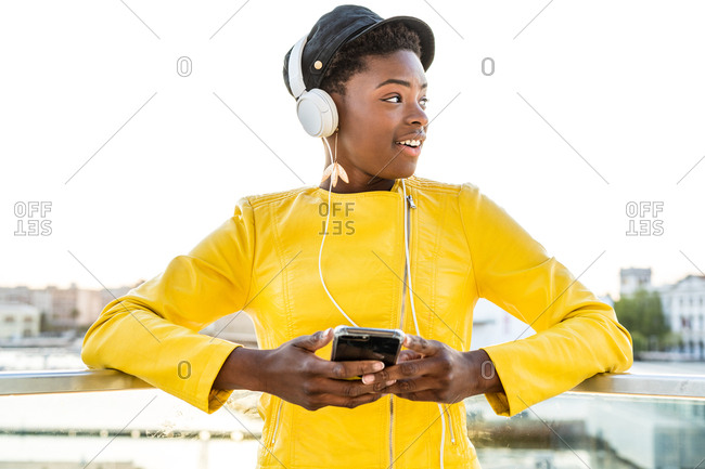 African American woman in stylish bright jacket using mobile phone and listen to music on headphones